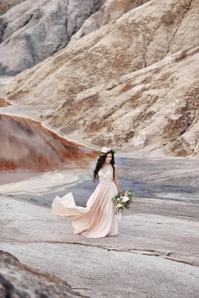 彼女の頭の上に花の花輪、手の美しい花の花束を持つ女性は、山の背景について説明します。長い光夏のドレスが風に揺れる. — ストック写真