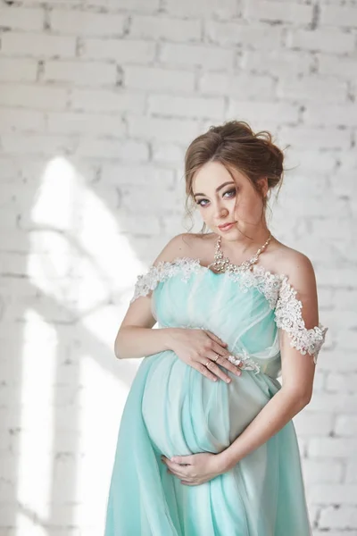 Mulher grávida bonita no vestido azul está esperando o nascimento de uma criança. O último mês de gravidez, mulher feliz. Mulher de mãos dadas sobre a barriga e posando no fundo da parede de tijolo — Fotografia de Stock