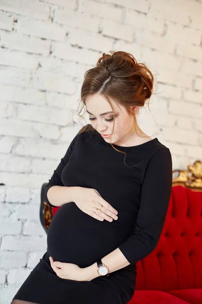 Όμορφη έγκυος γυναίκα κάθεται σε έναν κόκκινο καναπέ. Σε αναμονή για τη γέννηση του παιδιού. Εγκυμοσύνη — Φωτογραφία Αρχείου