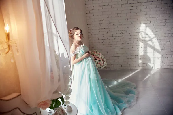 Беременная женщина стоит у окна в красивом лазурном платье. Портрет девочки, готовящейся стать матерью. Женщина в преддверии родов — стоковое фото