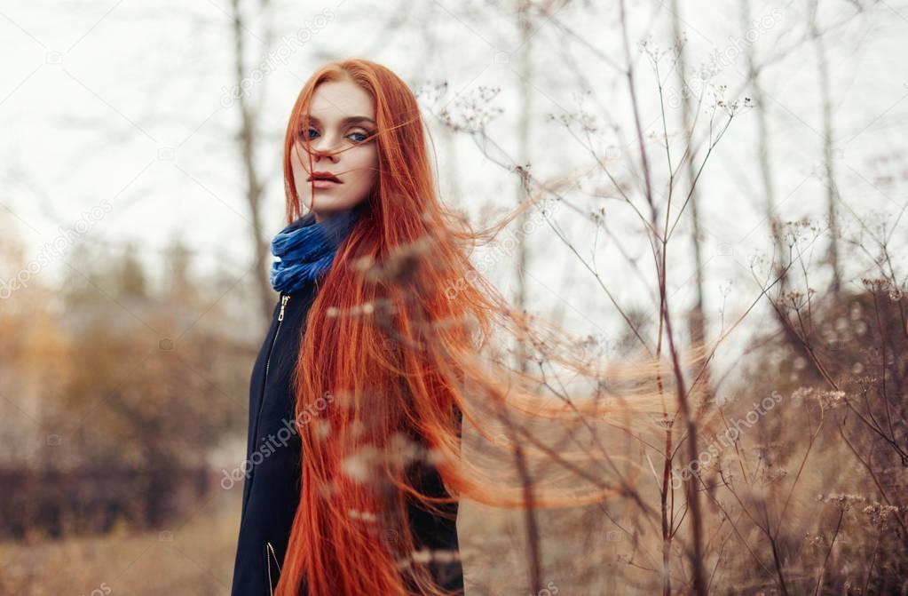 Woman with long red hair walks in autumn on the street. Mysterious dreamy look and the image of the girl. Redhead woman walking in the autumn the city. Cold cloudy autumn in the city.