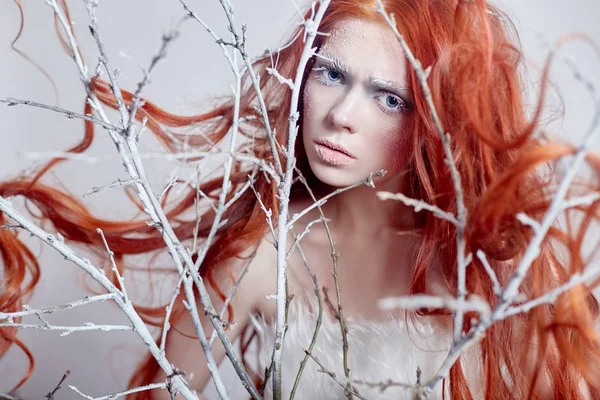 Redhead girl with long hair, a face covered with snow with frost. White eyebrows and eyelashes in frost, a tree branch covered with snow. Snow Queen and winter. Winter makeup woman face, red head.