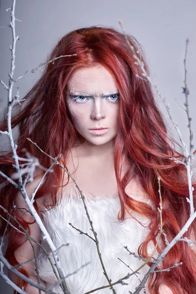 Руда дівчина з довгим волоссям, обличчя вкрите снігом з морозами. Білі брови та вії в мороз, гілка дерева вкрита снігом. Снігова Королева і зима. Зимовий макіяж обличчя жінки, червона голова . — стокове фото