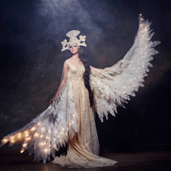 Арт-женщина ангел с крыльями в роскошном длинном платье и сказочный головной убор. Девушка-птица со светящимися крыльями позирует на темном фоне . — стоковое фото