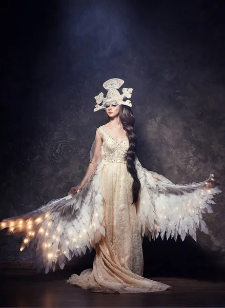 Арт-женщина ангел с крыльями в роскошном длинном платье и сказочный головной убор. Девушка-птица со светящимися крыльями позирует на темном фоне . — стоковое фото