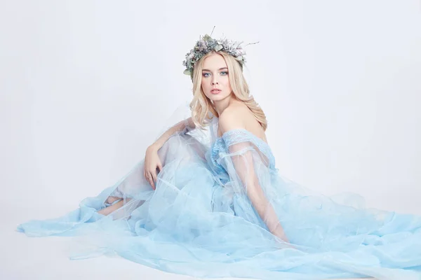 Charming fairy woman in a blue ethereal dress and a wreath on her head on white background, gentle mysterious blonde girl with perfect skin and makeup. Cleanliness, body care and skin.