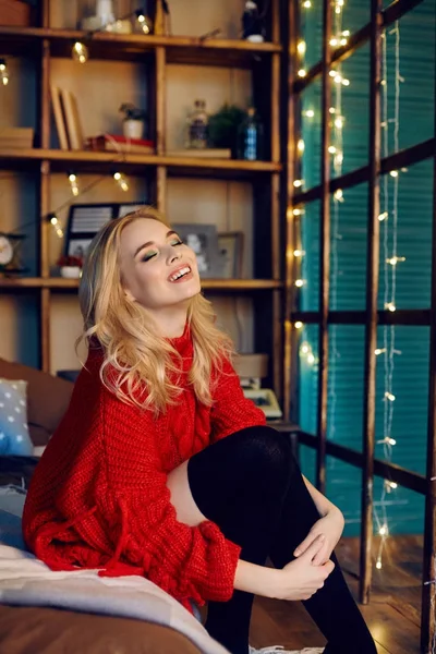 Sexig mode blond kvinna i röda tröjan sitter på sängen och ler. Glödande garland hemma. Porträtt av en flicka med en perfekt figur och ett leende. — Stockfoto
