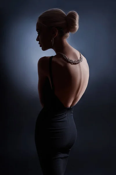 Ritratto donna di moda a contrasto su sfondo scuro, la silhouette di una ragazza con una bella schiena curva. La schiena nuda di una donna al buio. Lusso bionda posa in abito da sera . — Foto Stock