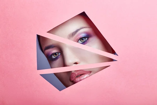 Kobieta z pięknie jasny makijaż i różowa szminka patrzy przez trójkątne szczelin w różowym papierze — Zdjęcie stockowe