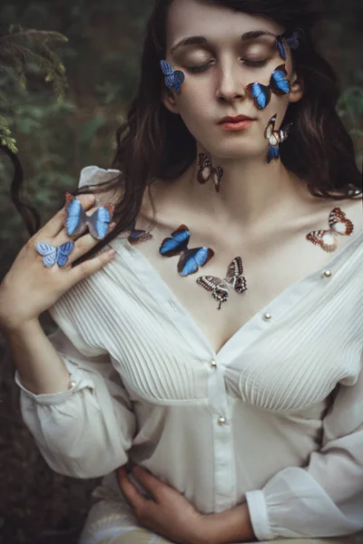 Bellissimo ritratto d'arte di una donna nuda con farfalle blu sul viso e le mani vicino al ramo di abete rosso nella natura nella foresta. Misteriosa magica romantica vista della ragazza vicino ai rami degli alberi . — Foto Stock