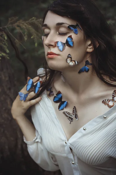 Hermoso retrato de arte de una mujer desnuda con mariposas azules en su cara y manos cerca de la rama de abeto en la naturaleza en el bosque. Misteriosa vista romántica mágica de la chica cerca de las ramas del árbol . —  Fotos de Stock
