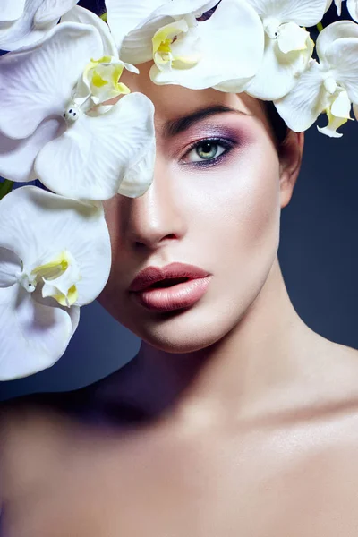 Chica morena con flores de orquídea en la cara y el pecho, retrato de belleza de un maquillaje perfecto, hermosos ojos y labios regordetes, jensina sobre un fondo púrpura oscuro. Cuidado de la piel facial y corporal . — Foto de Stock