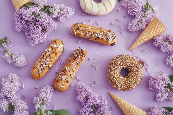 Donuts y pasteles de crema se encuentran sobre la mesa entre flores lila o — Foto de Stock