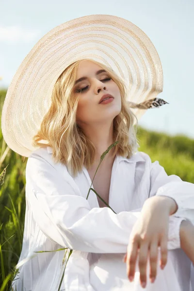 Ragazza con un grande cappello e un lungo vestito bianco è seduto nell'erba — Foto Stock