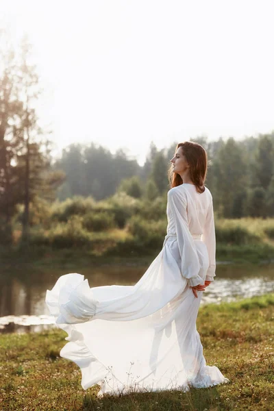 Beautiful slender woman in a long white dress walks in the morni