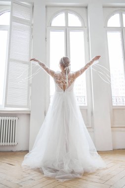 Büyük bir pencerenin yanında uzun beyaz bir elbise içinde, akşam güneşinde ince, güzel bir sarışın. Gelin için mükemmel saç ve makyaj, yeni bir gelinlik koleksiyonu.