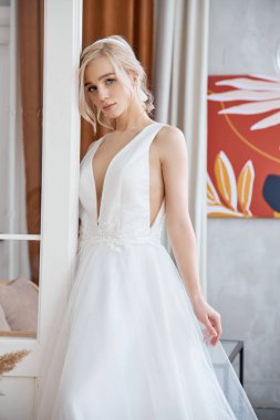 Büyük bir pencerenin yanında uzun beyaz bir elbise içinde, akşam güneşinde ince, güzel bir sarışın. Gelin için mükemmel saç ve makyaj, yeni bir gelinlik koleksiyonu.