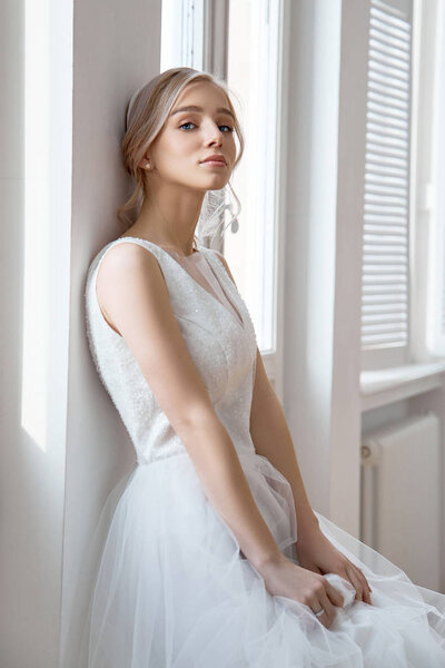 Невеста - это женщина в светлом летнем свадебном платье, стоящая у окна. Блондинка с прекрасными волосами и красивым макияжем
