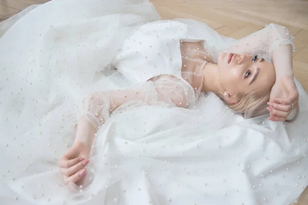 Romantic portrait of a woman on the floor in a beautiful long white dress. The girl is blonde with blue eyes and beautiful makeup on her face. Natural cosmetics
