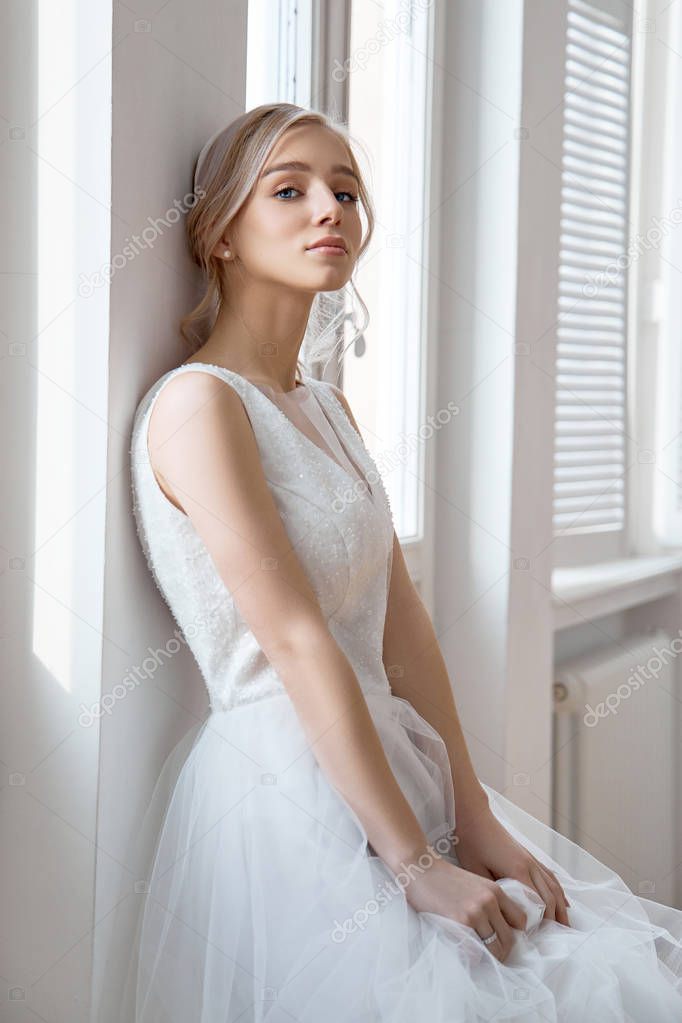 Bride is a woman in a light summer wedding dress standing at the window. Blonde girl with perfect hair and beautiful makeup