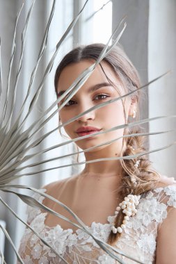 Gelin, pencerenin önünde duran açık renkli bir gelinlik giymiş bir kadın. Kahverengi saçlı, mükemmel saçlı ve güzel makyajlı bir kız.