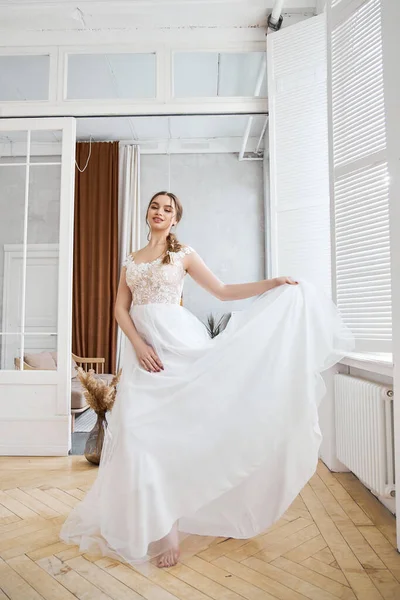 Bride is a woman in a light summer wedding dress standing at the window. Brown-haired girl with perfect hair and beautiful makeup
