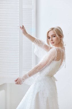 Evinde, pencerenin yanında güzel, uzun beyaz bir elbise giymiş bir kadının romantik portresi. Kız sarışın, mavi gözlü ve yüzünde güzel bir makyaj var. Doğal Kozmetik