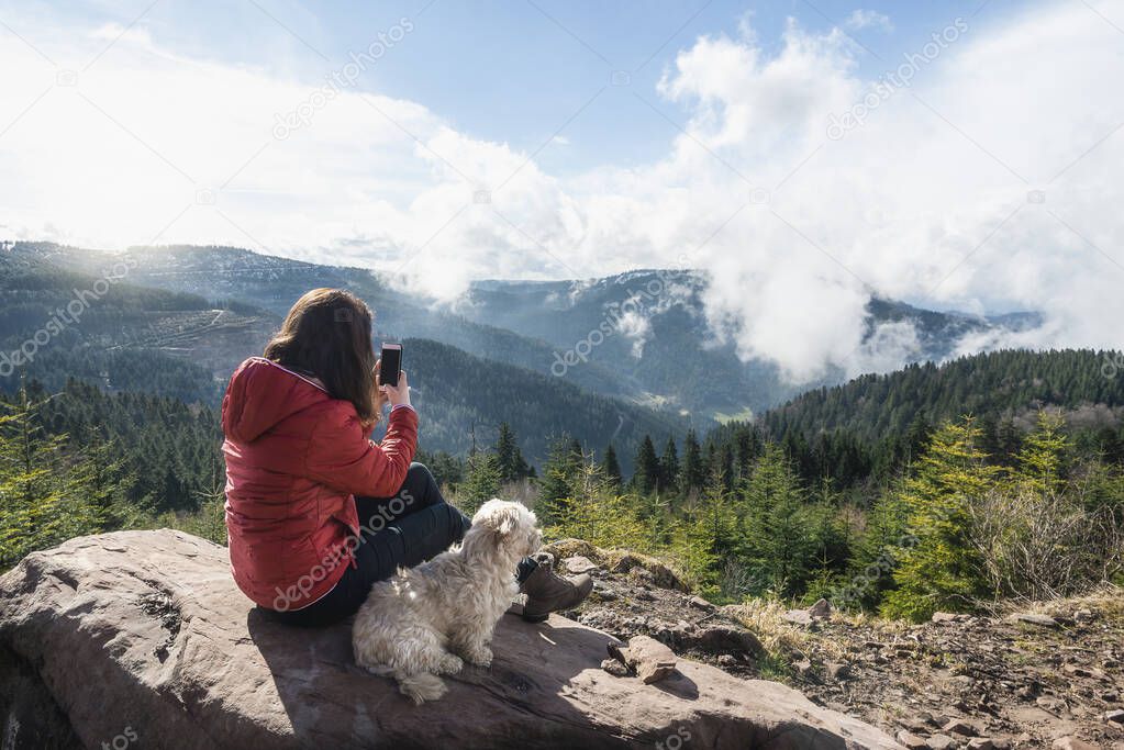Young woman with her dog admiring Black Forest mountains, on a sunny spring day, in Germany. Girl taking pictures with phone. Vacation time concept.