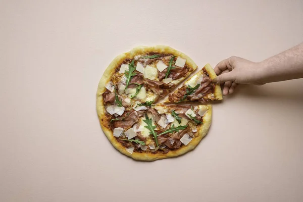 Sliced pizza prosciutto with arugula and cheese on pink background. Woman hand grabbing pizza slice top view. Italian traditional food.