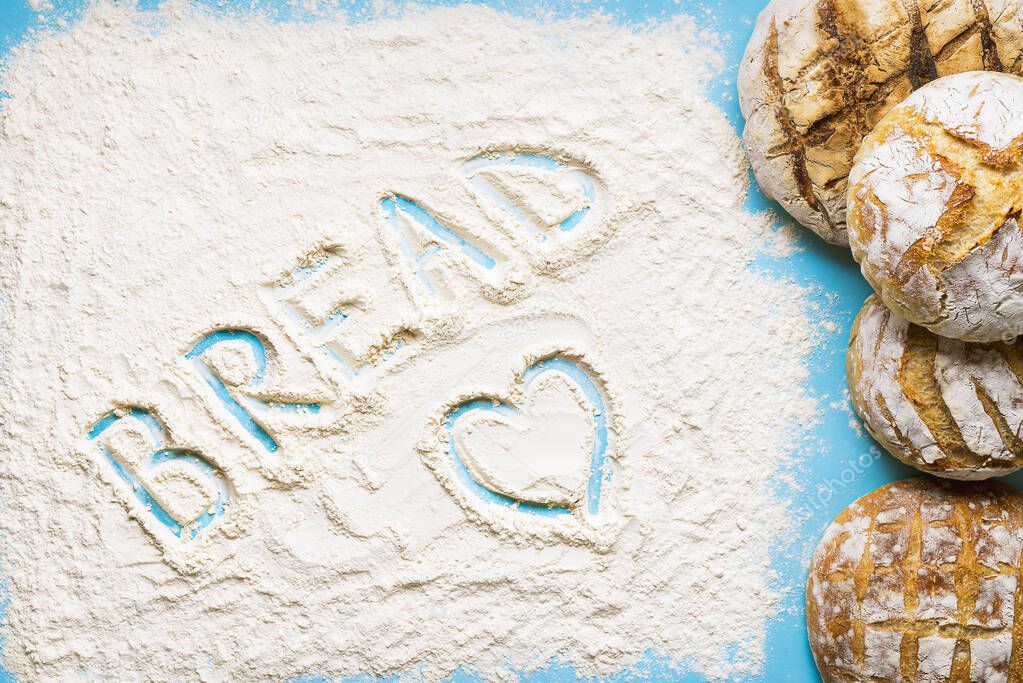 The word bread wrote in the flour spread on a blue table. Pile of round white loaves. Home-baked sourdough bread