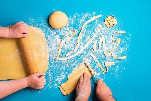 Couple preparing Italian pasta by hand on a blue background. Flat lay with making pasta. Rolling semolina yellow dough. Shaping tagliatelle pasta.