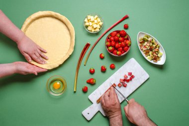 Yukarıda genç insanların elleri organik malzemelerle tatlı bir turta yapıyor. Çilek ve rhubarb keki, yeşil bir masanın üstünde, üst manzara