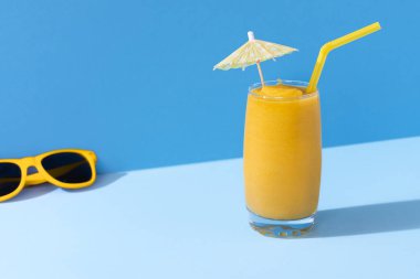 Mavi arka planda bir bardak mango smoothie ve sarı güneş gözlüğü, sert ışıkta. Güneşli yaz günü bağlamında. Tropik meyve püresi. Mango kokteyli.
