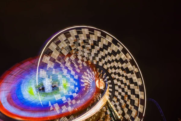 Dois passeios em movimento no parque de diversões, iluminação nocturna. Exposição longa . — Fotografia de Stock