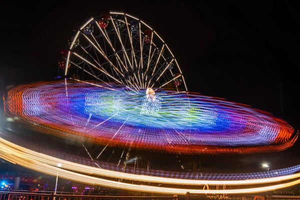 Dois passeios em movimento no parque de diversões, iluminação nocturna. Exposição longa . — Fotografia de Stock