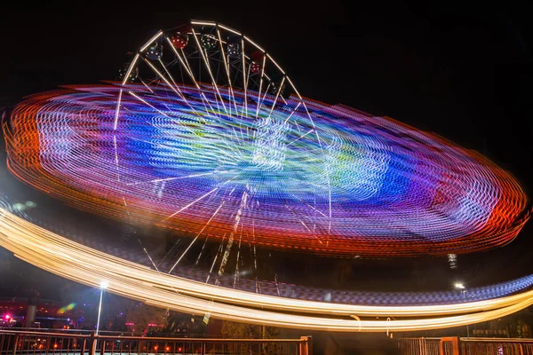 Due giostre in movimento nel parco divertimenti, illuminazione notturna. Lunga esposizione . — Foto Stock