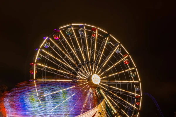 Due giostre in movimento nel parco divertimenti, illuminazione notturna. Lunga esposizione . — Foto Stock