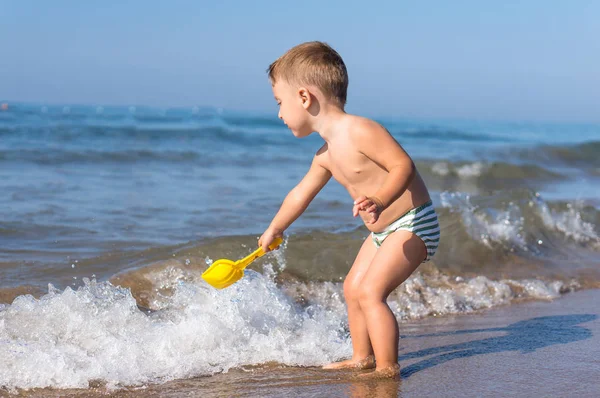 Kleiner Junge spielt mit gelber Spielzeugschaufel und winkt im Meer. — Stockfoto