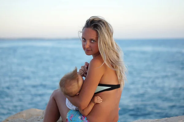 Mãe amamentando bebê na praia ao pôr do sol perto do mar — Fotografia de Stock