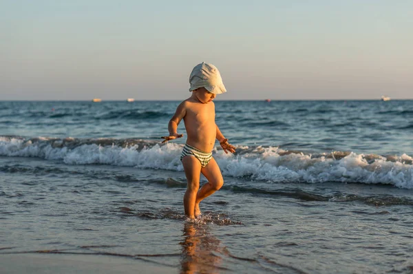 微笑小宝贝男孩在沙滩上在日落时运行时溅起水花在海. — 图库照片