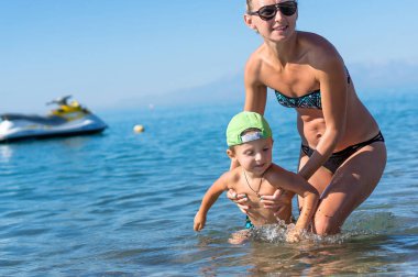 Genç anne siyah güneş gözlüğü ve denizde oyun gün süresi içinde yeşil Beyzbol şapkalı gülümseyen bebek çocuk evlat. Olumlu insan duyguları, duygular, sevinç.