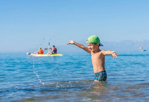 Усміхнений маленький хлопчик в зеленій бейсбольній шапці грає і бризкає в морі, океані. Позитивні людські емоції, почуття, радість. Бабуся і дідусь катаються на каяках у тропічному океані вдень . — стокове фото