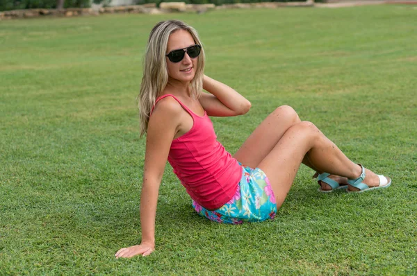 Atractivo joven sonriente hermoso pelo rubio rizado delgada chica retrato de moda en rosa camiseta posando mirando lejos mirada tranquila, en el prado de hierba verde de fondo. Concepto de estilo de vida saludable mujer. Emociones humanas positivas, sentimientos, alegría. Gafas de sol negras —  Fotos de Stock