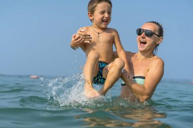 Genç anne siyah güneş gözlüğü ve denizde oyun gün süresi içinde yeşil Beyzbol şapkalı gülümseyen bebek çocuk evlat. Olumlu insan duyguları, duygular, joy. Bahar ve yaz tatil.