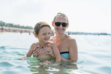 Genç anne siyah güneş gözlüğü ve denizde oyun gün süresi içinde gülümseyen bebek çocuk evlat. Olumlu insan duyguları, duygular, joy. Sıçrama. Bahar ve yaz tatil.