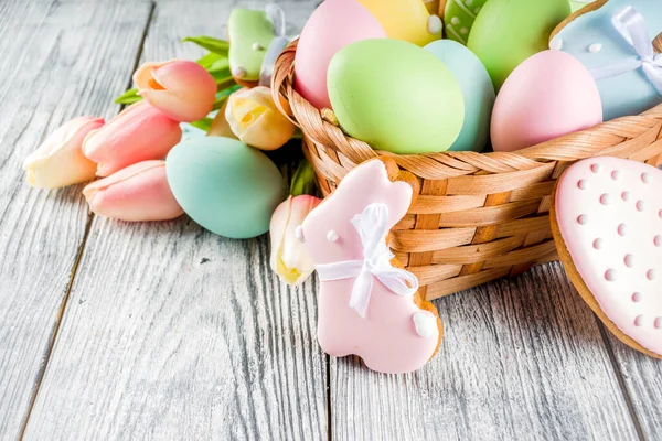 Easter greeting card background with pastel colored eggs and homemade cookies shaped in eggs and bunnies rabbits. With a basket, tulips, rustic wooden table, copy space