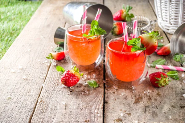 Red summer drink with ice, cold strawberry mojito or lemonade with fresh strawberries and mint on sunlight summer wooden and glass background, copy space