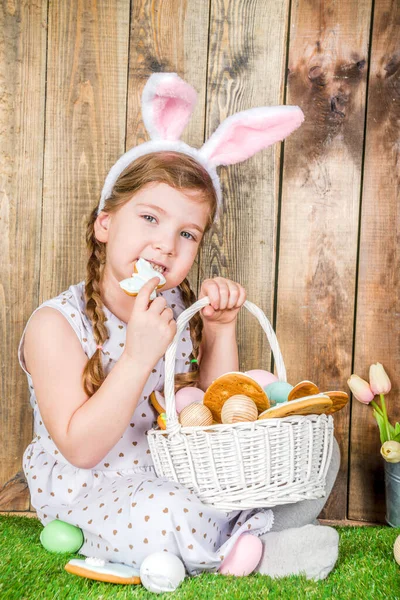 Cute little child girl with bunny ears on Easter holiday. With painted eggs basket, bunny toy and sweet Easter cookies. Easter greeting card background, wooden background with green spring lawn.