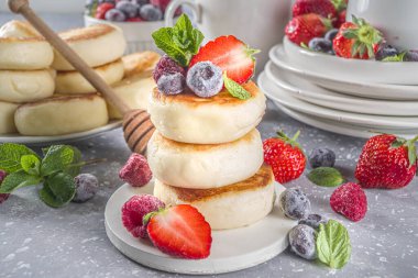 Bal sürülmüş süzme peynir krepleri ve gri arka planda çeşitli yaz meyveleri, yaz kahvaltısı veya öğle yemeği