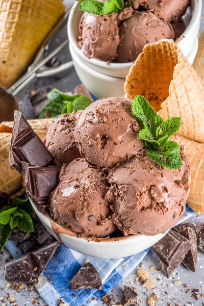 Homemade chocolate ice cream with chocolate pieces and shavings, and ice cream cones. In small white bowls on white grey stone table copy space